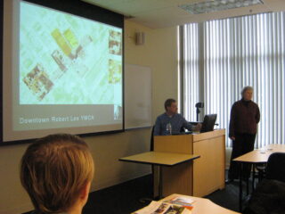 A man stands at the front of the room in front of a screen giving a presentation to a group of people.