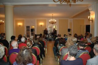 Two men sit in chairs at the front of a room, in front of an audience.