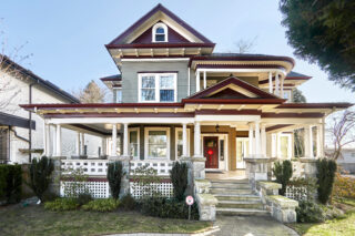 A recently renovated large home featuring style markers from Edwardian, Victorian and Queen Anne architecture. The house features a small staircase leading to the entryway in a large porch area, with a front facing bay-window and cylindric columns holding the next level of this 2 1/2 storey home. The main floor's roofline features a front-gabled roof covering the entryway and upholding a circular balcony with similar columns. There is fretwork throughout the roof skirt of this home. To the right of the balcony there is another front-gabled roof piece with a dormer for the half upper floor.