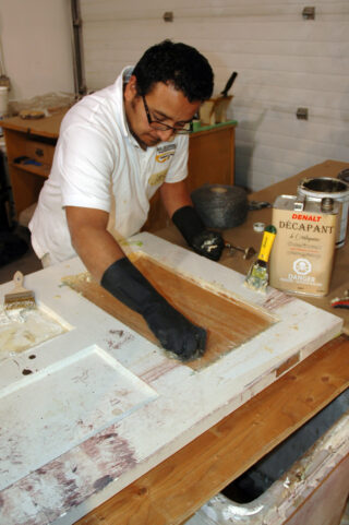 A man is removing white paint from the middle of a wood door.