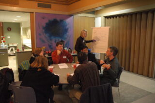 Group of people sitting around a table discussing and one woman writes on white chart paper with a marker.