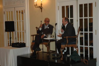 Two men sit in chairs holding microphones, talking.