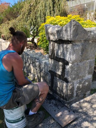 A mason sitting on a bucket, repointing a stone pillar