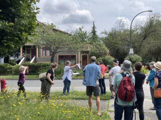 A woman stands in front of a heritage house, holding up a piece of paper to show to the group.