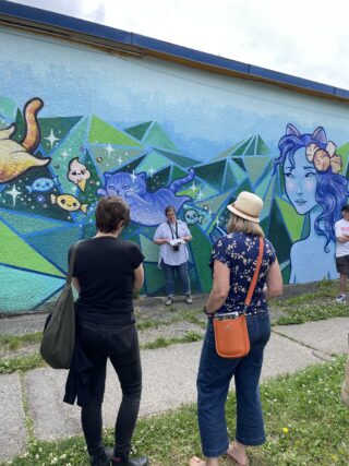 A woman stands in front of a colourful mural, talking to a walking tour group.